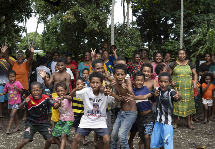 Children in Papua New Guinea pose for the camera while their families are in the background by trees.