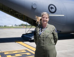 A woman stands on the tarmac smiling at the camera, her blond ponytail blowing in the wind while she wears a green flying suit with the New Zealand patch on her left arm.  A large grey aircraft is behind her, the ramp is down and the New Zealand Kiwi is o