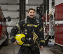 A firefighter stands in uniform, with a helmet in his hand, between two fire trucks in a fire station.