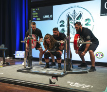 Corporal Nicole Smith on stage in front of a large screen ready to powerlift large weights while three spotters are on hand behind her.