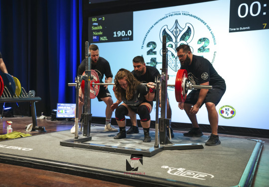 Corporal Nicole Smith on stage in front of a large screen ready to powerlift large weights while three spotters are on hand behind her.