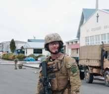 A man stands wearing camouflage uniform, including a helmet, vest and weapon stands on a road in Stratford. In the background a tan-coloured Army vehicle and buildings of local businesses.
