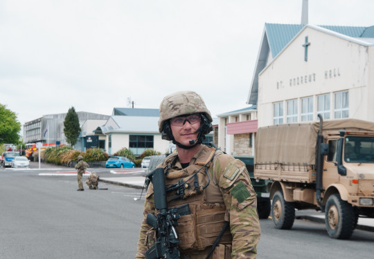 A man stands wearing camouflage uniform, including a helmet, vest and weapon stands on a road in Stratford. In the background a tan-coloured Army vehicle and buildings of local businesses.