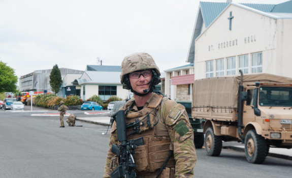 A man stands wearing camouflage uniform, including a helmet, vest and weapon stands on a road in Stratford. In the background a tan-coloured Army vehicle and buildings of local businesses.
