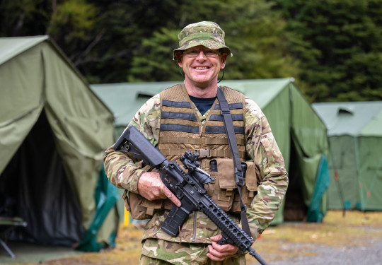 An aviator in camouflage holding a MARS-L smiles at the camera in front of large dark green tents. 