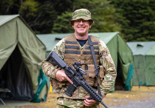 An aviator in camouflage holding a MARS-L smiles at the camera in front of large dark green tents. 
