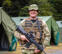An aviator in camouflage holding a MARS-L smiles at the camera in front of large dark green tents. 
