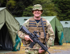 An aviator in camouflage holding a MARS-L smiles at the camera in front of large dark green tents. 
