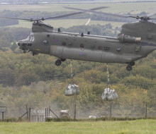 A Chinook helicopter delivers two large bags of chalk to the green hillside in Wiltshire. A gate in the foreground reads 'The Kiwi'.