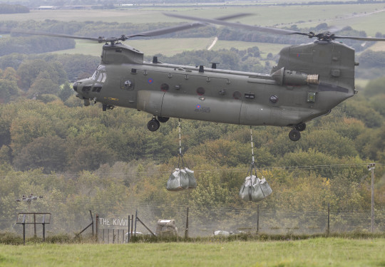 A Chinook helicopter delivers two large bags of chalk to the green hillside in Wiltshire. A gate in the foreground reads 'The Kiwi'.