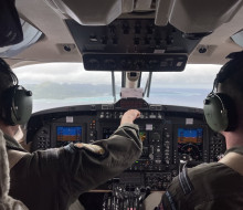 Two people in the flight deck of an aircraft, looking out the window. One of them is touching something on the panels. 