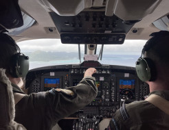 Two people in the flight deck of an aircraft, looking out the window. One of them is touching something on the panels. 