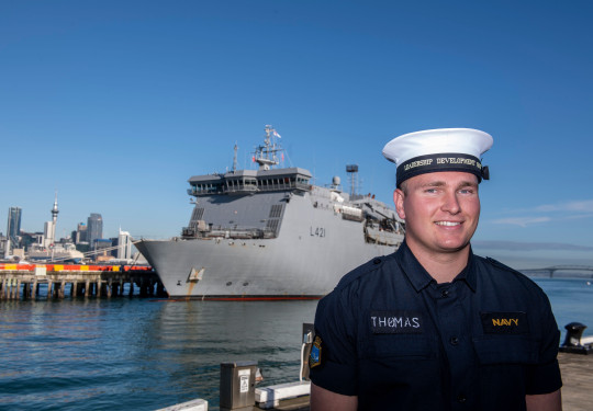 A sailor stands at Devonport Naval Base with HMNZS Canterbury in the background on a sunny day, blue sky