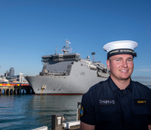 A sailor stands at Devonport Naval Base with HMNZS Canterbury in the background on a sunny day, blue sky