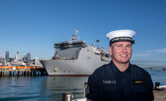 A sailor stands at Devonport Naval Base with HMNZS Canterbury in the background on a sunny day, blue sky