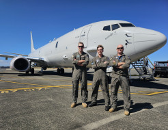 Three aviators in Air Force uniform stand with arms crossed in front of a large grey jet-powered aircraft under blue skies.