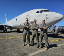 Three aviators in Air Force uniform stand with arms crossed in front of a large grey jet-powered aircraft under blue skies.