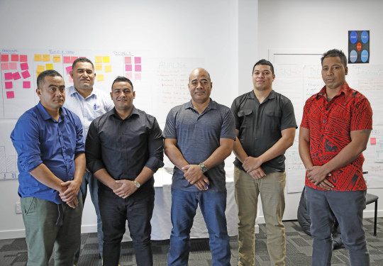 A group of people stand in a line for the photo in a classroom with brainstorming post-it notes in the background