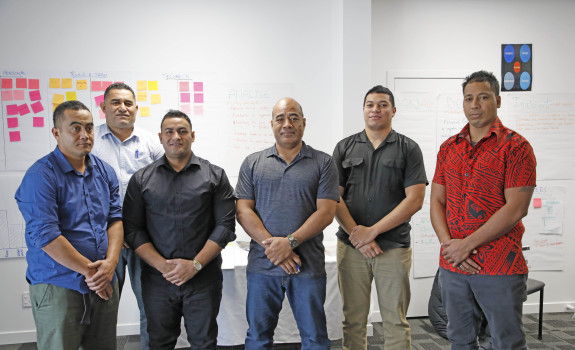 A group of people stand in a line for the photo in a classroom with brainstorming post-it notes in the background