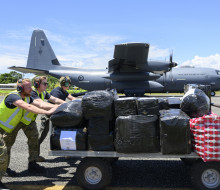 Three people push a trolley full of cargo in front of a large grey aircraft.