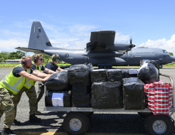 Three people push a trolley full of cargo in front of a large grey aircraft.