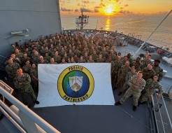 A badge printed on a sign which reads, "Pacific Partnership" is held at the front of a large group of personnel at the from of a military ship. In the background the sun sets, reflecting off the water and lighting the sky and clouds.