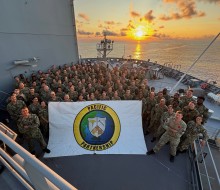 A badge printed on a sign which reads, "Pacific Partnership" is held at the front of a large group of personnel at the from of a military ship. In the background the sun sets, reflecting off the water and lighting the sky and clouds.