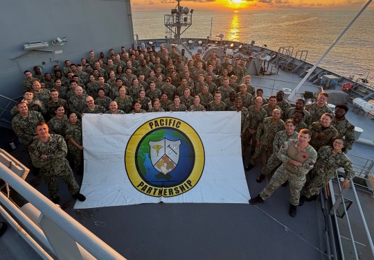 A badge printed on a sign which reads, "Pacific Partnership" is held at the front of a large group of personnel at the from of a military ship. In the background the sun sets, reflecting off the water and lighting the sky and clouds.
