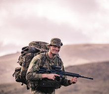 A soldier during early morning pack march, holding a weapon and smiling at the camera