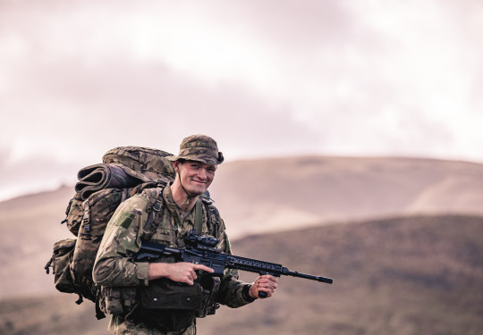 A soldier during early morning pack march, holding a weapon and smiling at the camera
