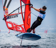 Corporal Haybittle on his windfoil, a blue board with a mat travelling down to the water and a red and white kite on top. He is pulling the shaka and is wearing a black helmet, glasses and wetsuit.