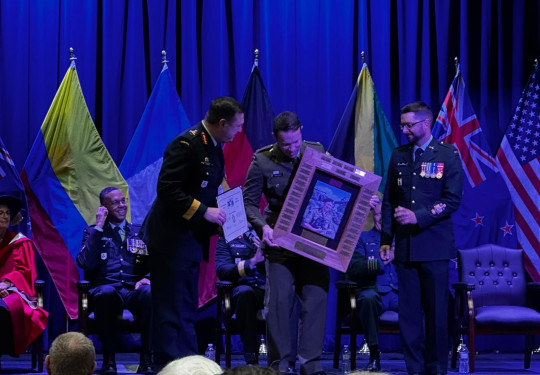 Major Prouse recieved a trophy on stage from two other military personnel. The lighting is very blue and flags line the back of the stage in front of a stage curtain.
