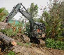 A bulldozer sits atop mud as it pushes a pile of tree debris to the side. Several workers wearing high vis safety gear work in the background.