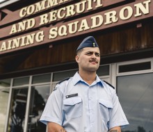A male wearing an Air Force light blue shirt and a blue cap stands outside of a building with the words 'Command and Recruit Training Squadron'
