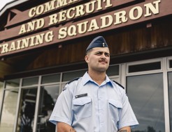 A male wearing an Air Force light blue shirt and a blue cap stands outside of a building with the words 'Command and Recruit Training Squadron'