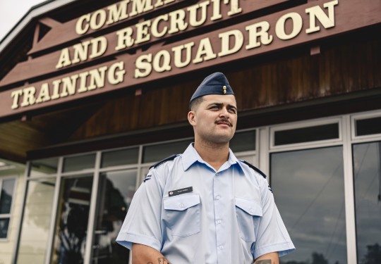 A male wearing an Air Force light blue shirt and a blue cap stands outside of a building with the words 'Command and Recruit Training Squadron'