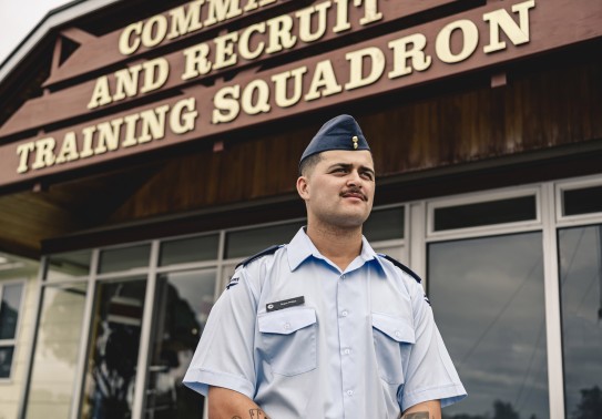 A male wearing an Air Force light blue shirt and a blue cap stands outside of a building with the words 'Command and Recruit Training Squadron'