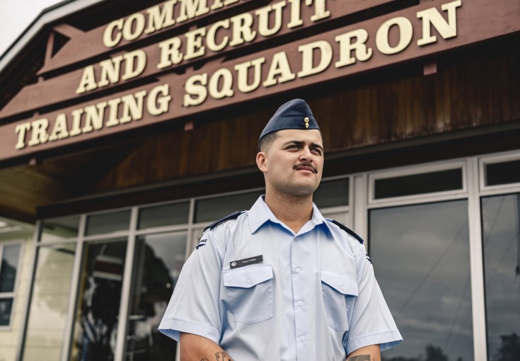 A male wearing an Air Force light blue shirt and a blue cap stands outside of a building with the words 'Command and Recruit Training Squadron'