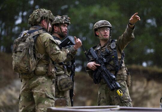 Three soldiers in Army uniform holding weapons having a discussion. The solider on the right points into the distance.