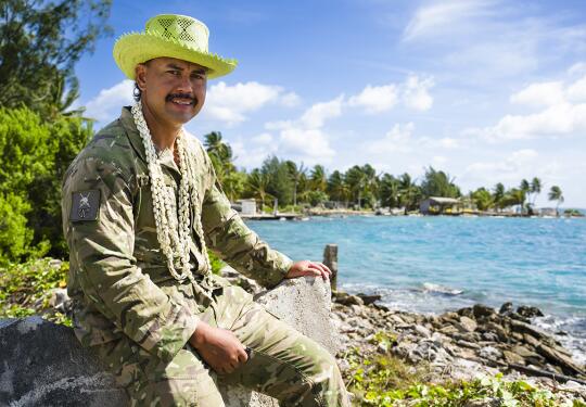 Person in camouflage uniform wearing a bright hat and shell garland seated on a concrete wall beside a tropical shoreline with turquoise water and palm trees.