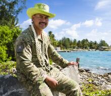 Person in camouflage uniform wearing a bright hat and shell garland seated on a concrete wall beside a tropical shoreline with turquoise water and palm trees.