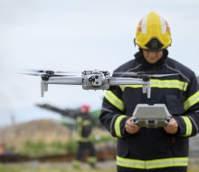 A drone flies with its four rotors spinning in front of a firefighter who is operating the drone from a handheld console.  The male firefighter wears a black jacket with high visibility panels, he looks down at the console and wears a yellow helmet.  In t