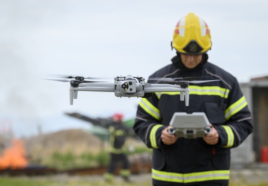 A drone flies with its four rotors spinning in front of a firefighter who is operating the drone from a handheld console.  The male firefighter wears a black jacket with high visibility panels, he looks down at the console and wears a yellow helmet.  In t