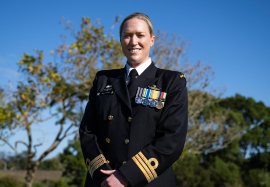 A woman in dress Navy uniform smiles at the camera in front of trees and blue sky.