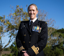 A woman in dress Navy uniform smiles at the camera in front of trees and blue sky.