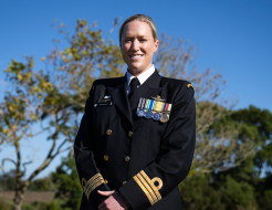 A woman in dress Navy uniform smiles at the camera in front of trees and blue sky.