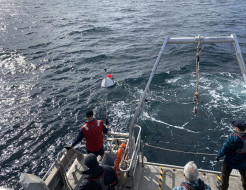 A floating white reflector a slightly smaller fluorescent pink bouy in the sea, seen from a ship just off its stern. Four people can be seen from behind looking towards the reflector, one is holding a boat hook.