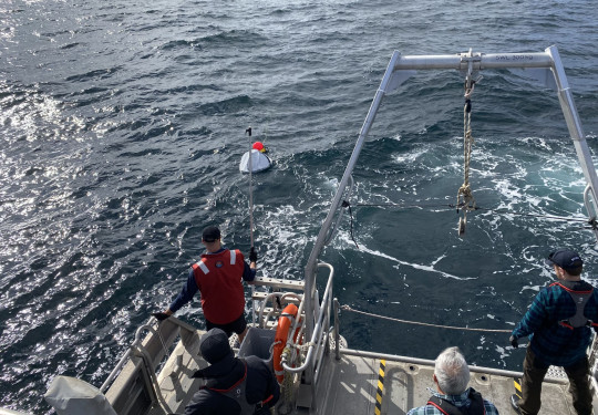 A floating white reflector a slightly smaller fluorescent pink bouy in the sea, seen from a ship just off its stern. Four people can be seen from behind looking towards the reflector, one is holding a boat hook.