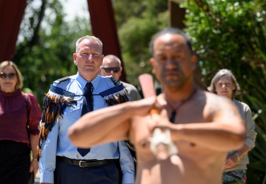 Peter Gibson makes his way onto the Turangawaewae wearing a korowai. He is being lead onto the marae by a toa (warrior) who is welding a taiaha (wooden staff)