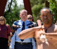 Peter Gibson makes his way onto the Turangawaewae wearing a korowai. He is being lead onto the marae by a toa (warrior) who is welding a taiaha (wooden staff)